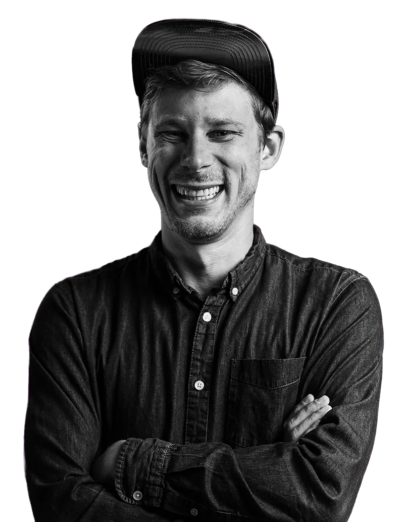 Ein lächelnder Mann in schwarzem Hemd mit Kappe vor lila Hintergrund, schwarz-weiß Porträt.