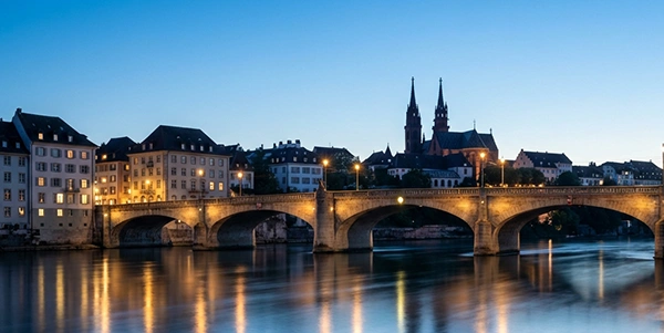 Abendliche Skyline von Basel mit beleuchteter Brücke und typischen Gebäuden am Rhein im sanften Dämmerlicht.