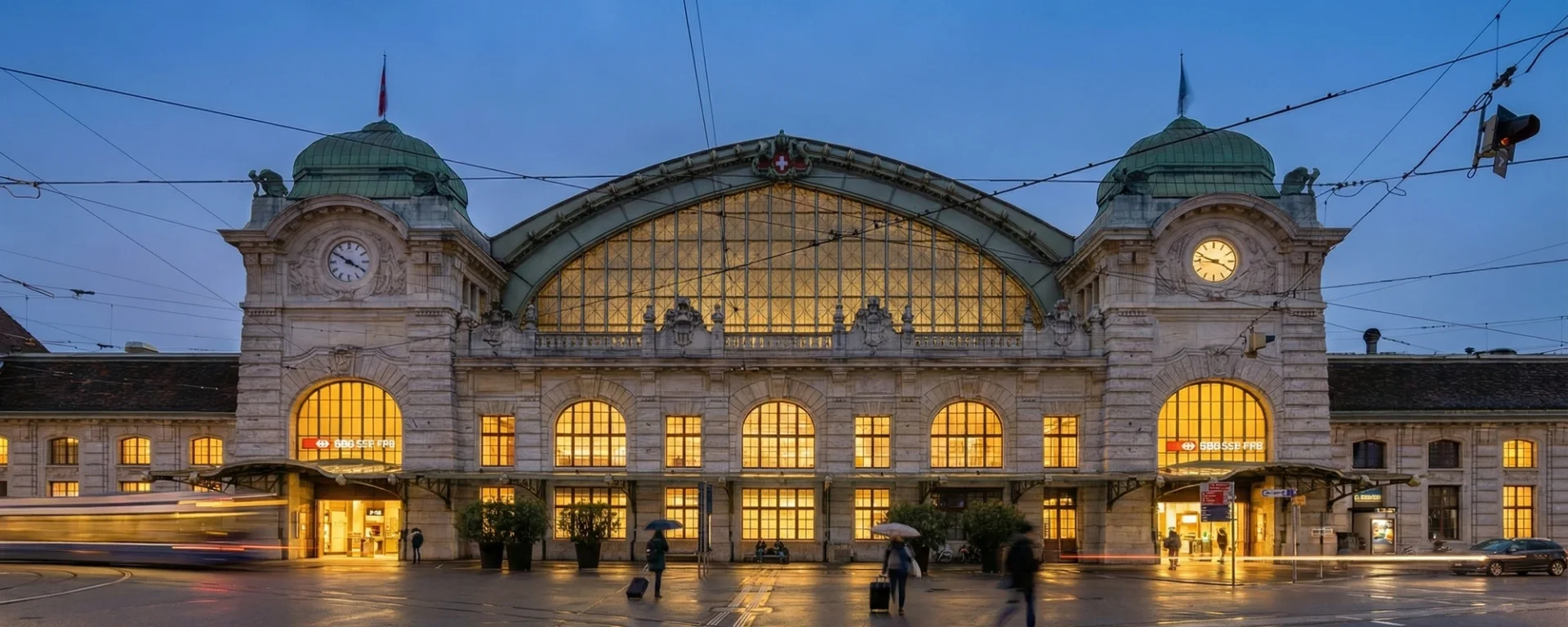 Bahnhof SBB in Basel bei Dämmerung mit beleuchteten Fenstern und Uhrtürmen.