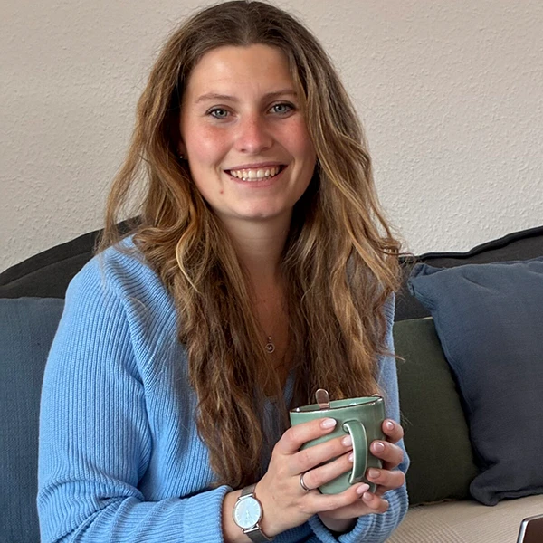 Frau in blauem Pullover sitzt auf Sofa und hält eine Tasse, lächelnd in gemütlicher Wohnzimmeratmosphäre.