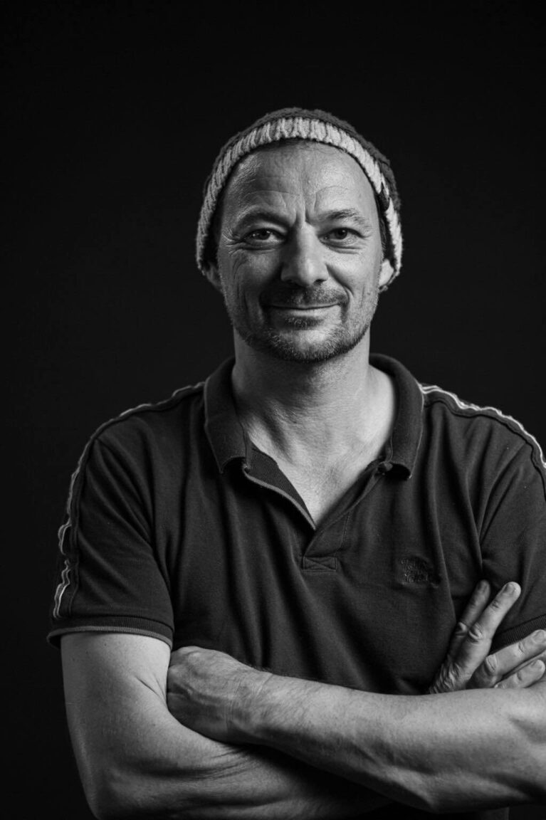 Schwarzweißporträt eines lächelnden Mannes mit Mütze und verschränkten Armen vor neutralem Hintergrund.