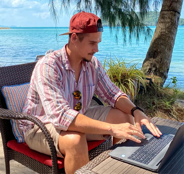 Mann arbeitet am Laptop in tropischer Kulisse am Strand, mit Palmen und Meer.