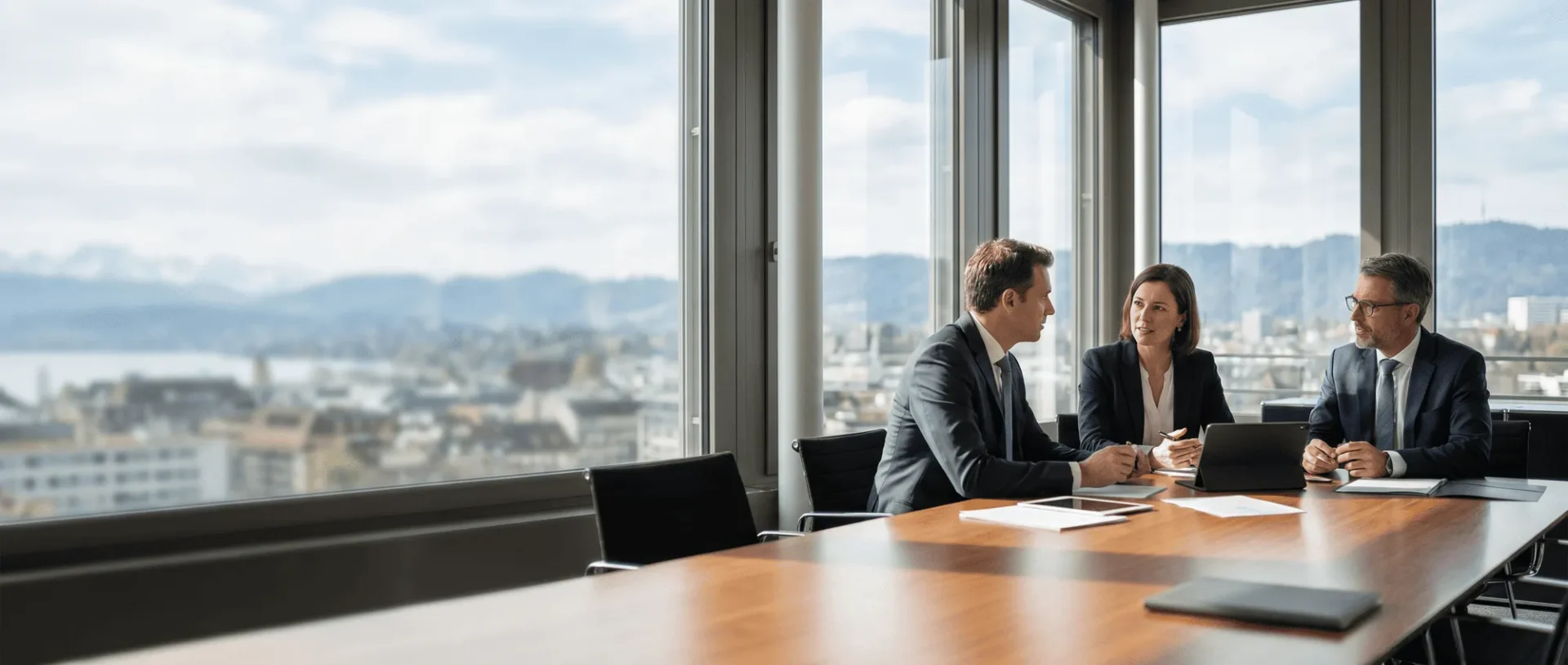 Geschäftstreffen in modernem Büro mit Panoramafenster, drei Personen diskutieren am Konferenztisch; Stadtlandschaft im Hintergrund.