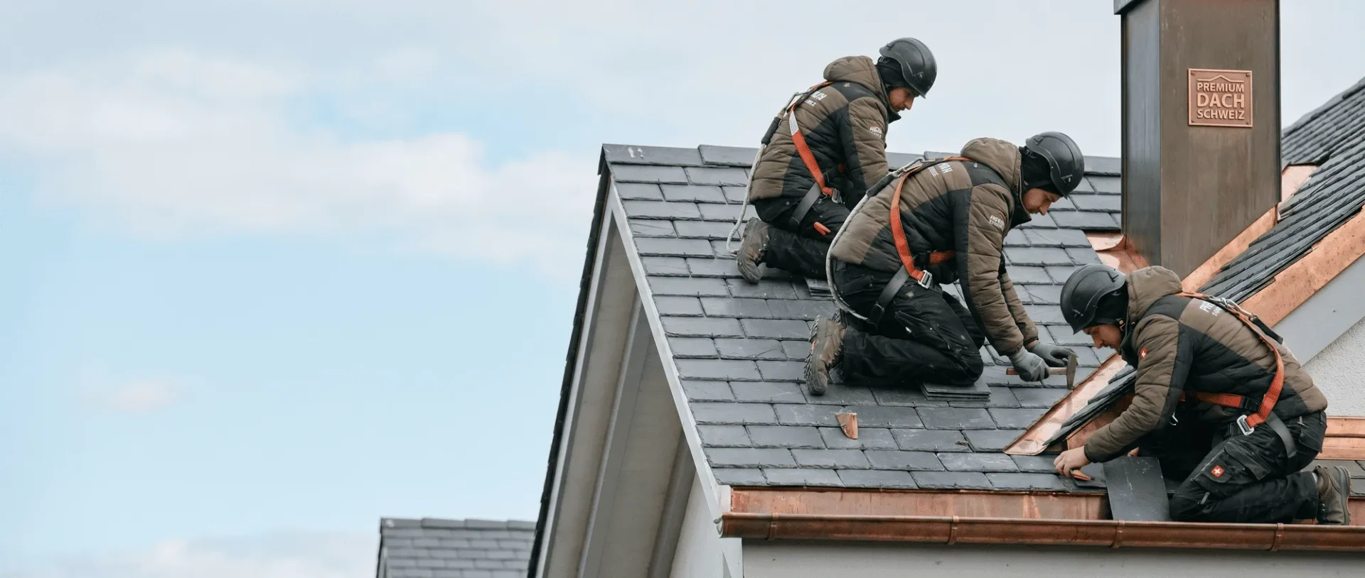 Handwerker reparieren Dachziegel auf einem Schieferdach bei klarem Himmel, ausgestattet mit Sicherheitsausrüstung.