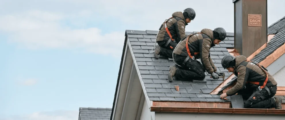 Handwerker reparieren Dachziegel auf einem Schieferdach bei klarem Himmel, ausgestattet mit Sicherheitsausrüstung.